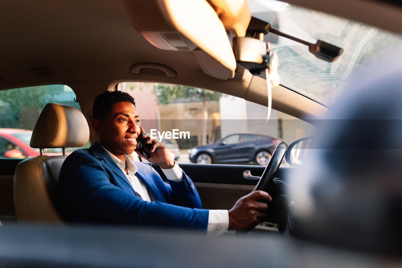 Side view of successful young african american male entrepreneur in elegant formal suit talking on mobile phone while driving car on city street