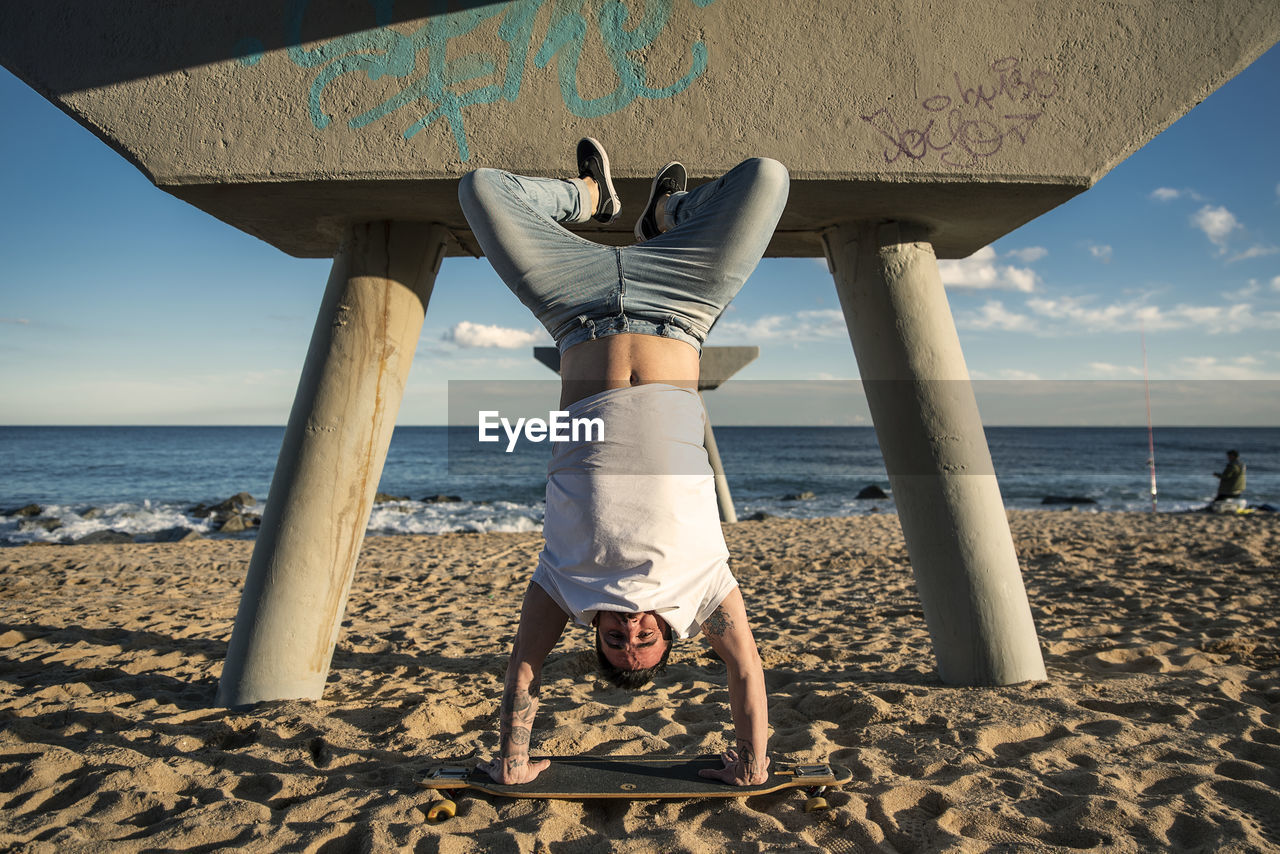 Mid adult man doing headstand on surfboard at beach against sky during sunny day