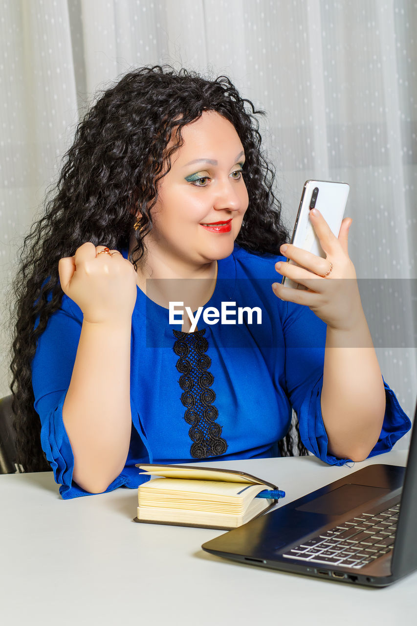 Curly brunette woman sits at a table in the office working with a smartphone. vertical photo