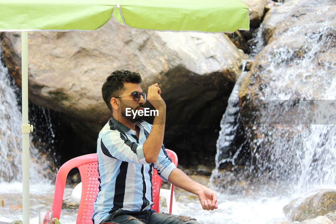 Young man sitting on chair against waterfall