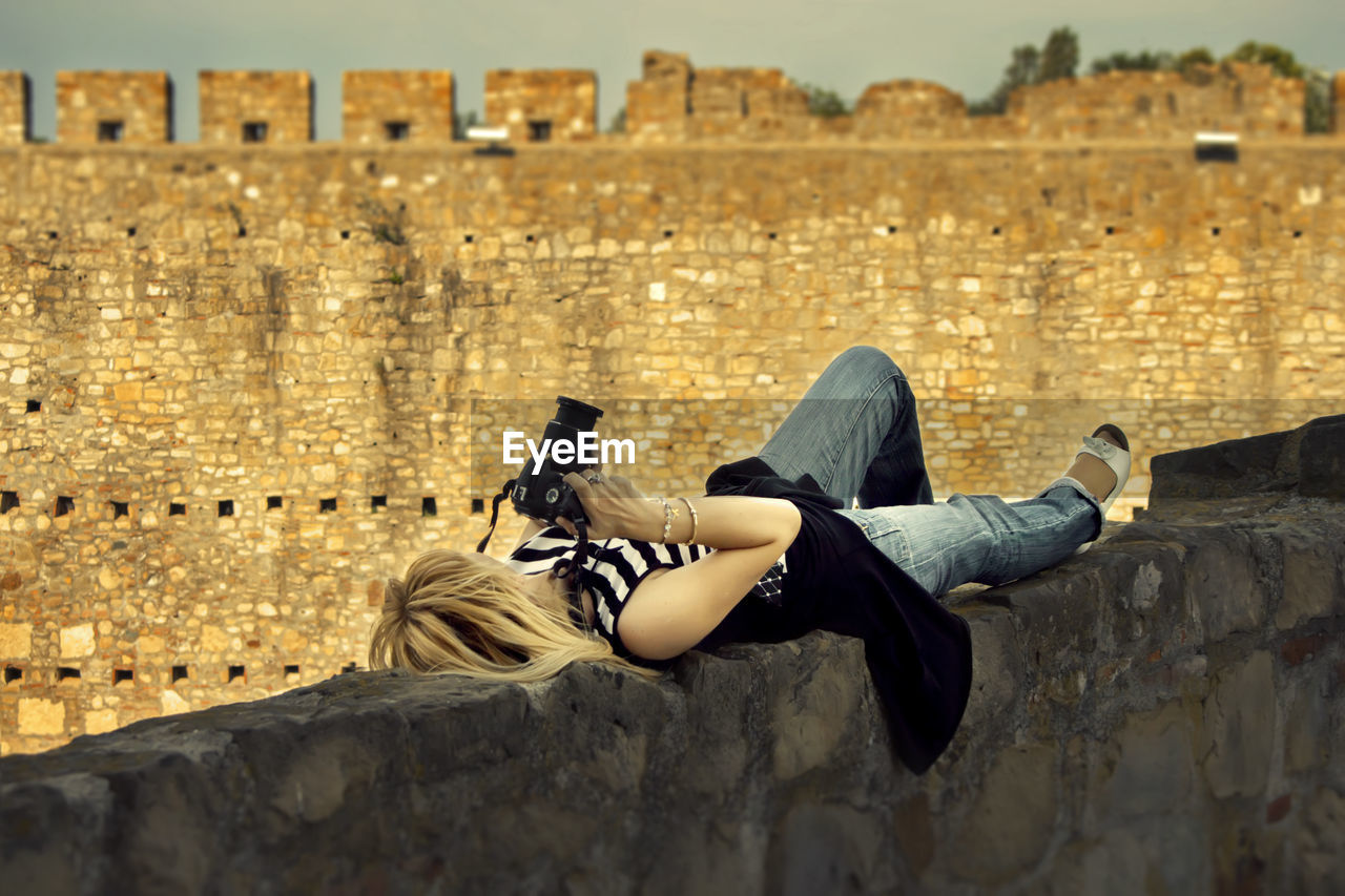 Mature woman with camera lying on retaining wall against old ruin