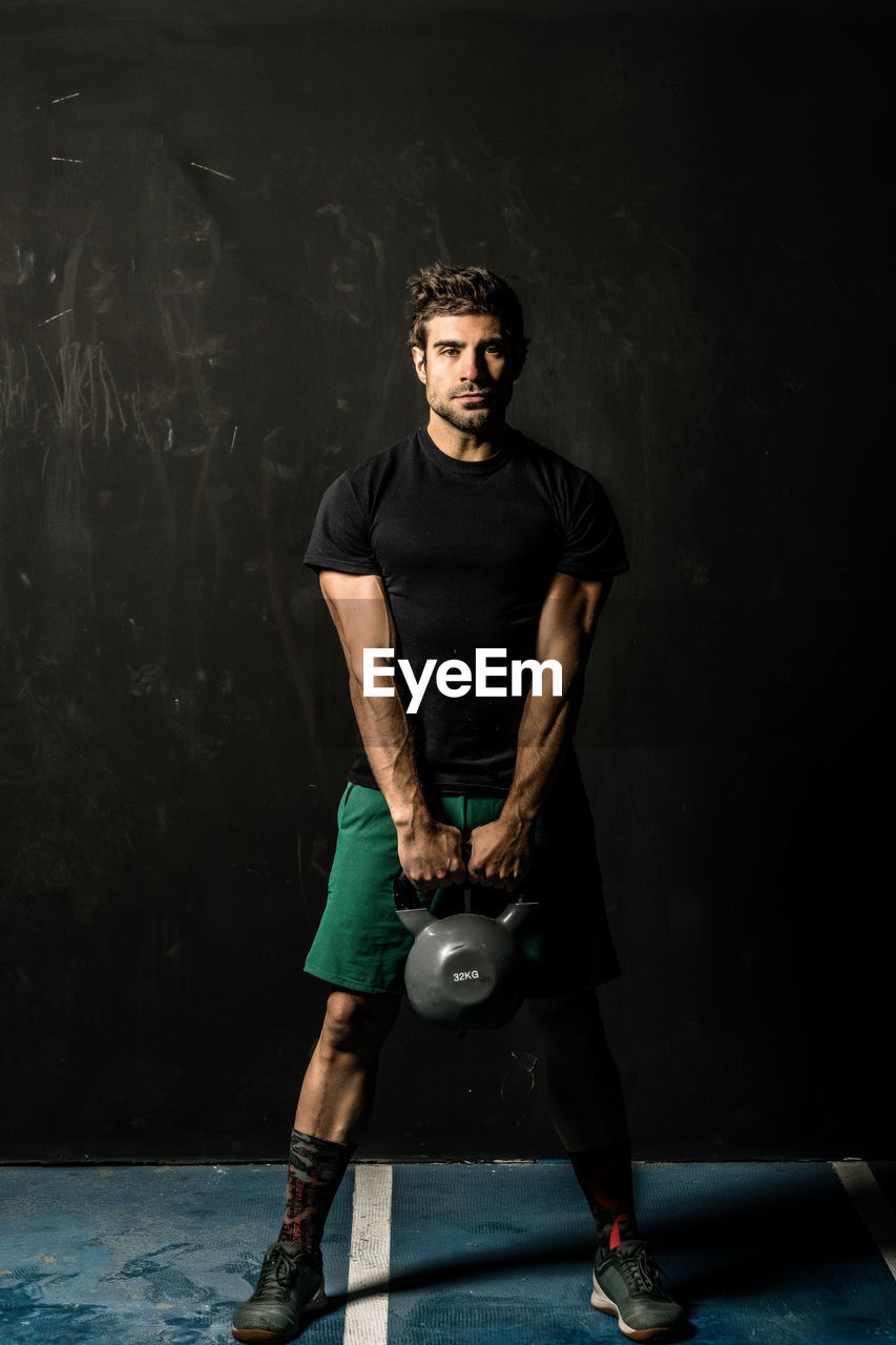 Full length powerful male athlete lifting heavy kettlebell while standing against black wall during functional workout in gym