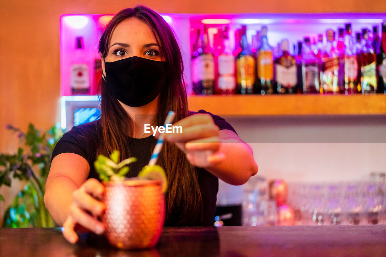 Young female barkeeper in protective mask serving cocktail with straw while working in bar