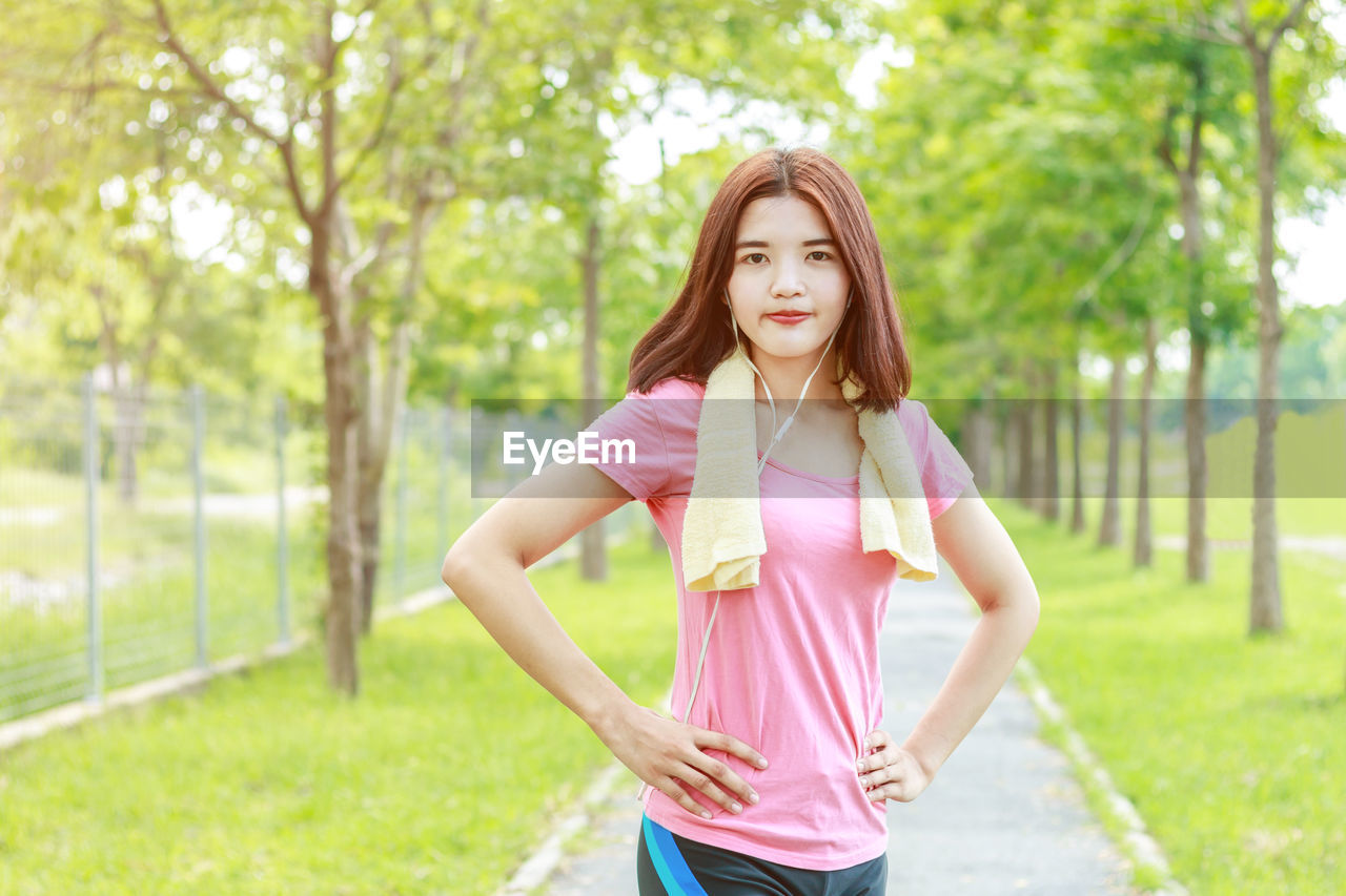 Portrait of woman listening music while standing on footpath against trees