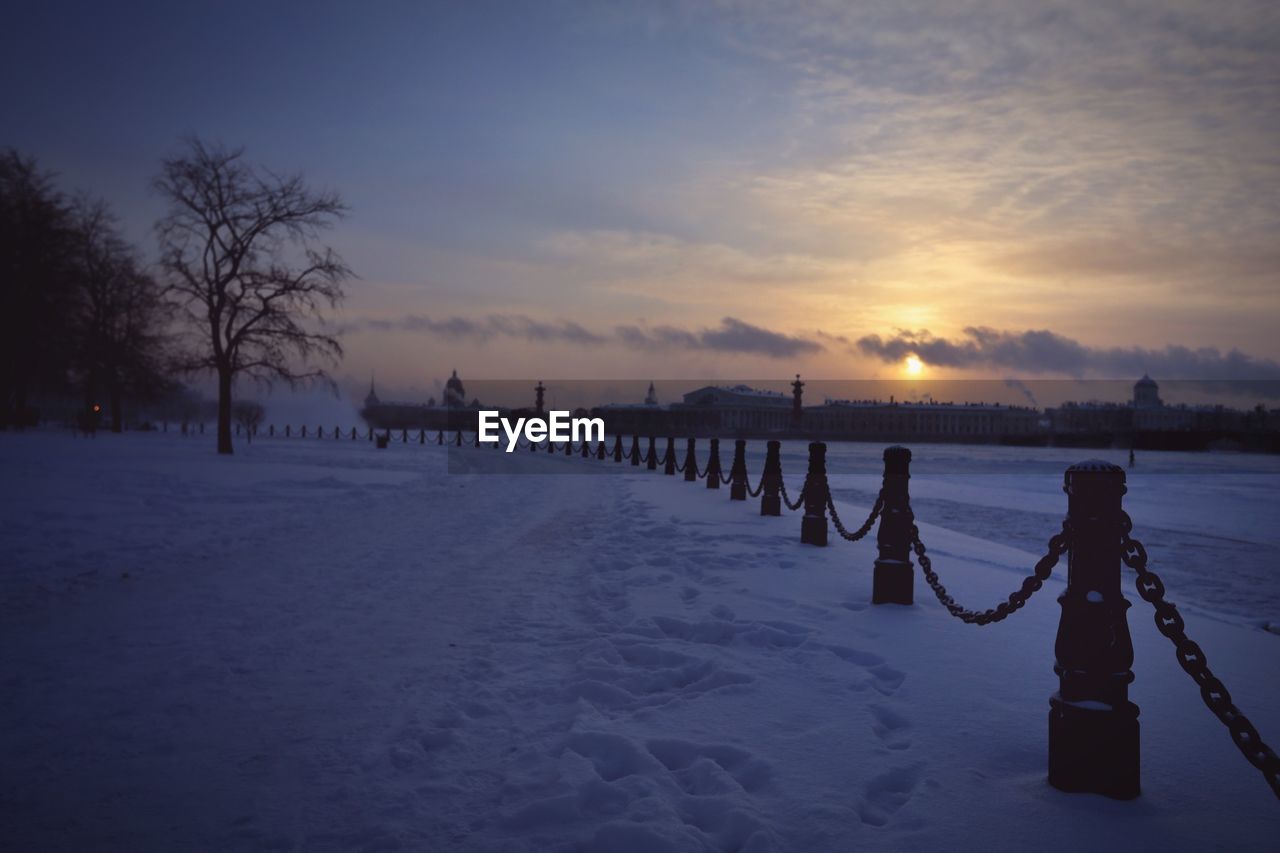 Railing on snow covered field against sky during sunset