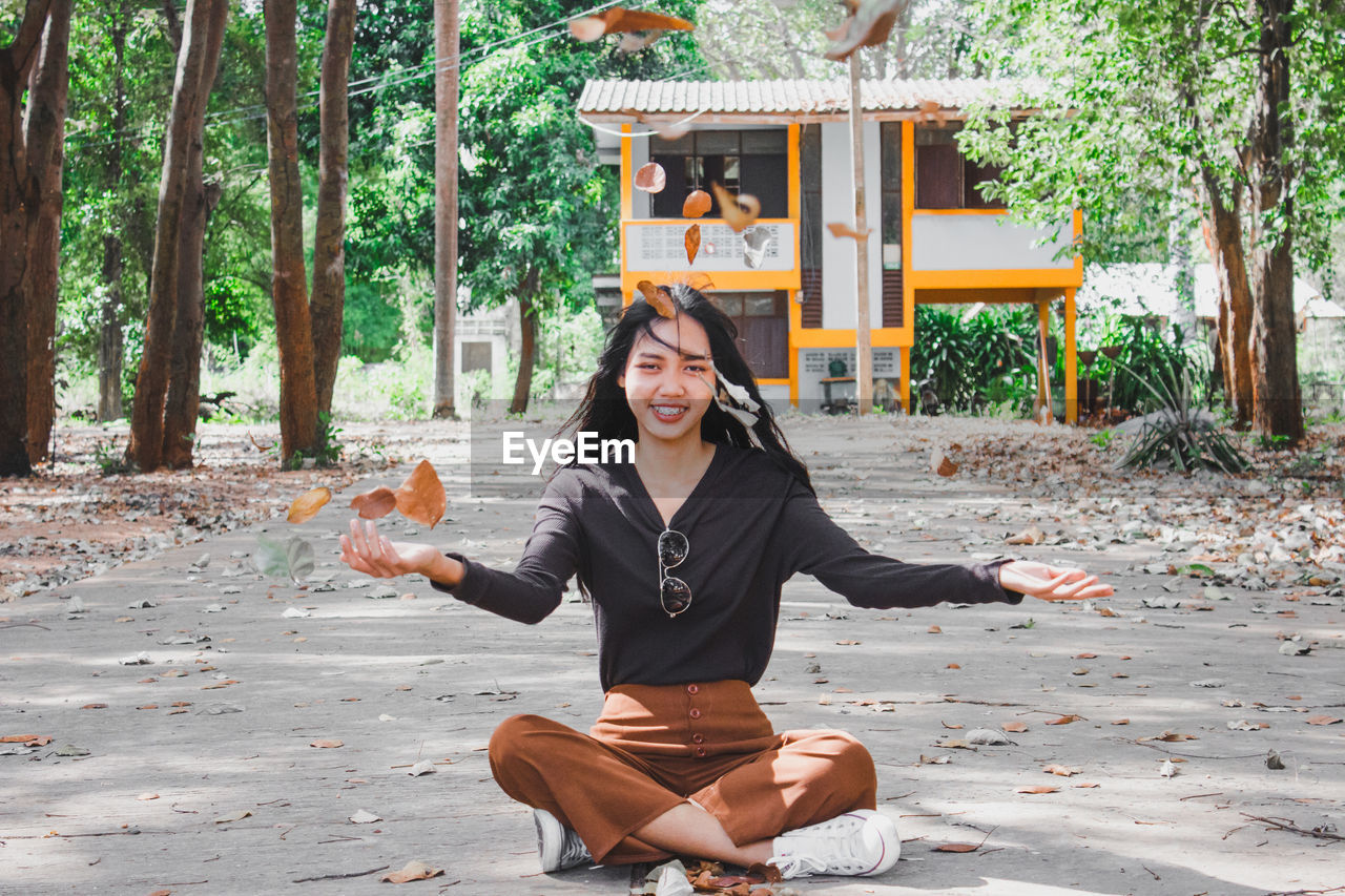 Portrait of smiling young woman throwing leaves while sitting on road in forest