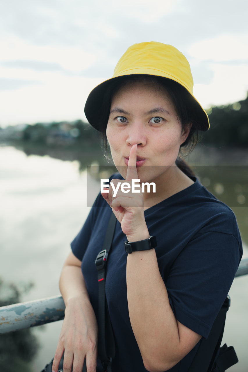 Portrait of woman with finger on lips wearing hat while standing by railing over canal against sky
