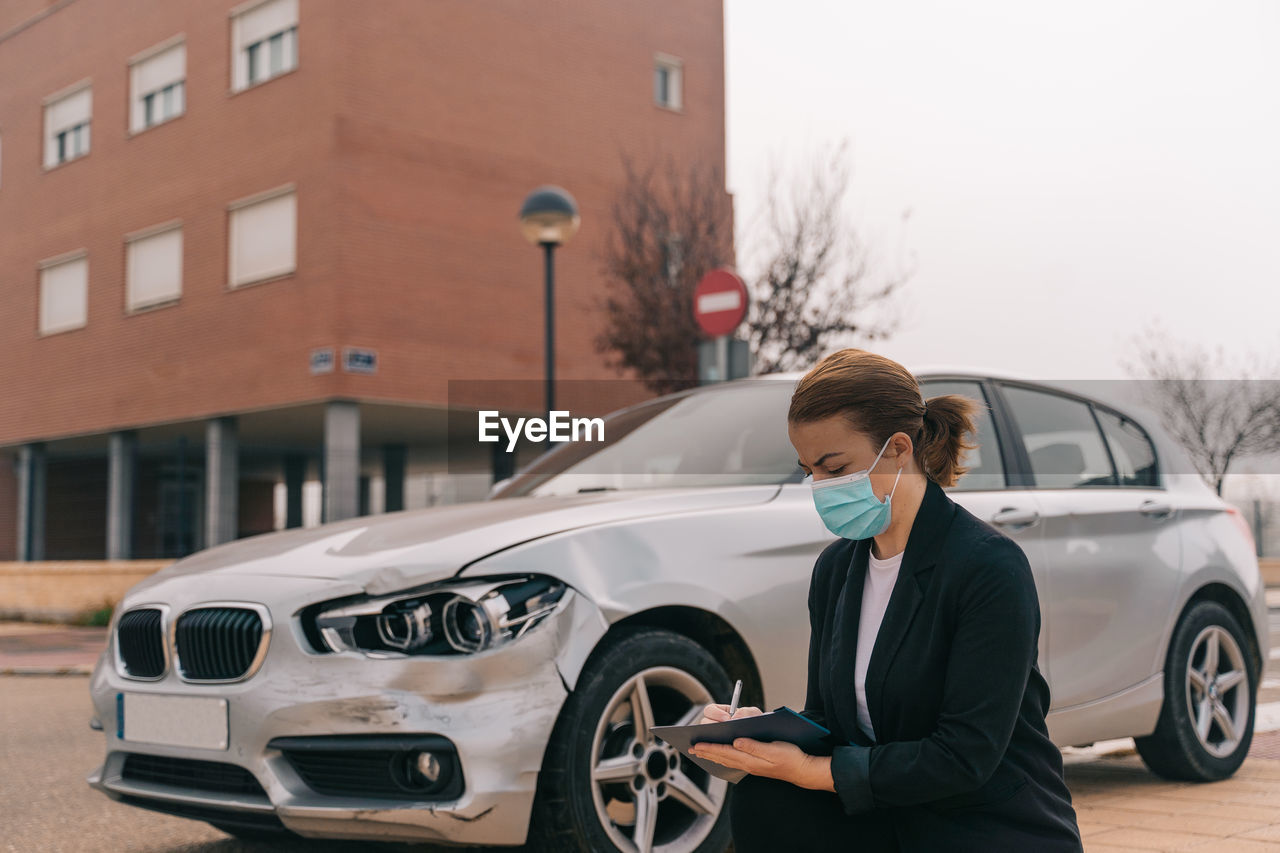 Side view of unrecognizable female insurance agent in formal outfit and protective mask writing protocol near damaged car parked on street after accident