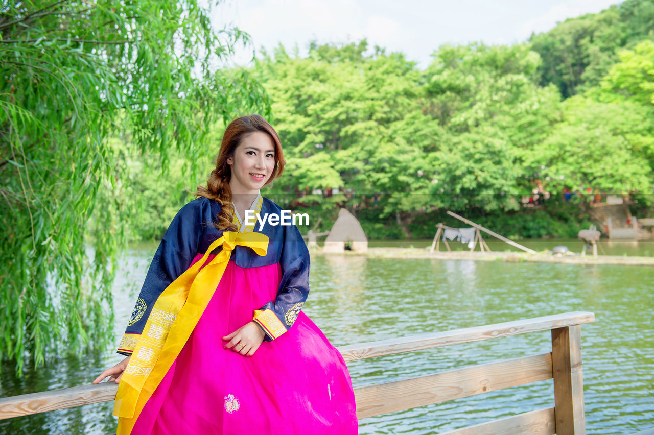 Portrait of smiling beautiful woman wearing hanbok while sitting on railing against lake