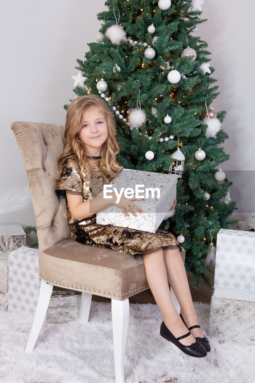 Full length of girl sitting on chair by christmas tree at home
