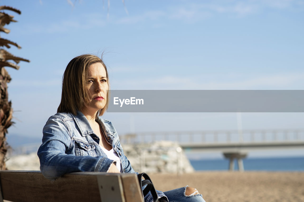 Thoughtful woman sitting on a bench near sea while looking away