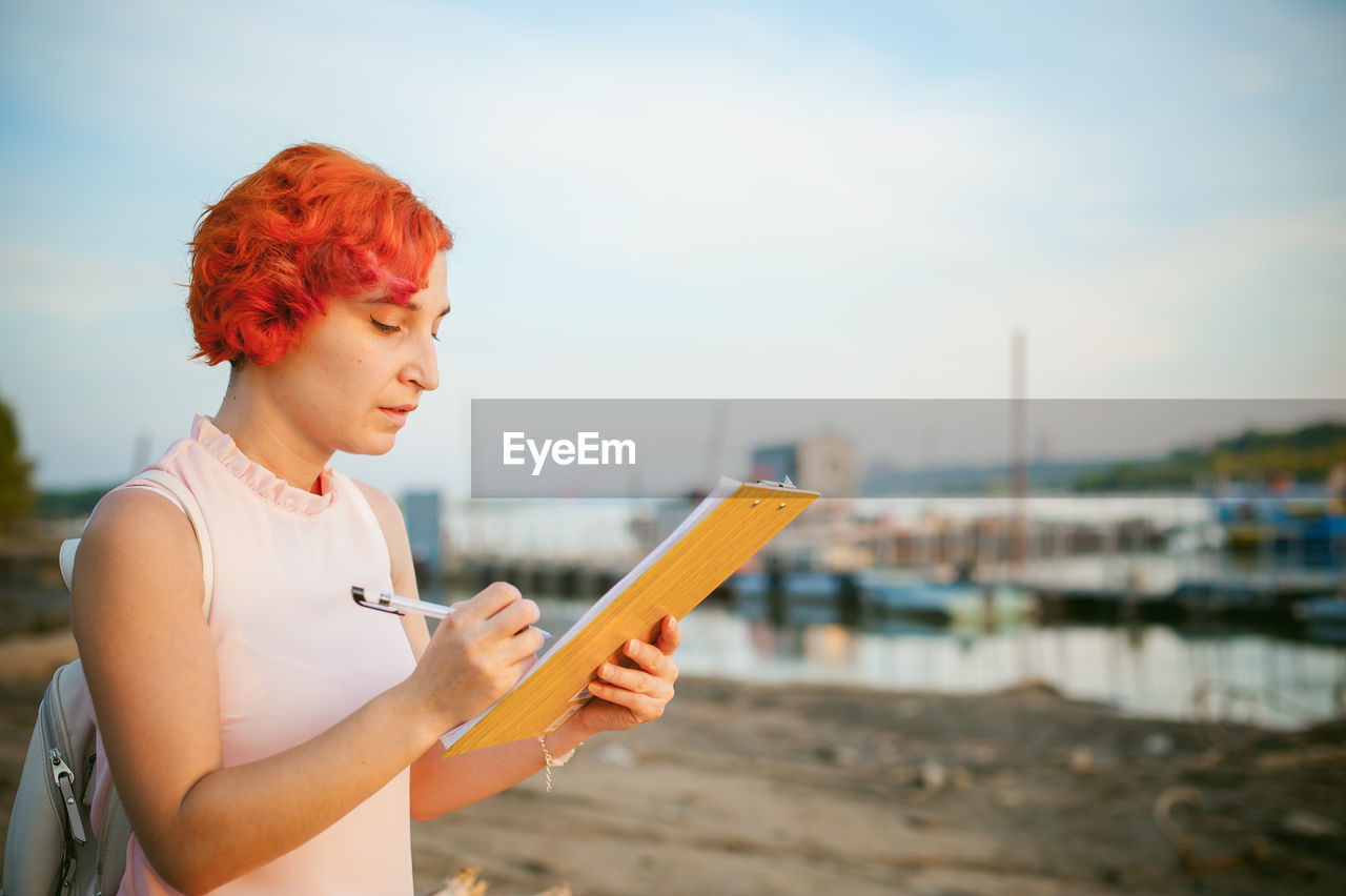 Mid adult woman writing while standing at riverbank against sky during sunset