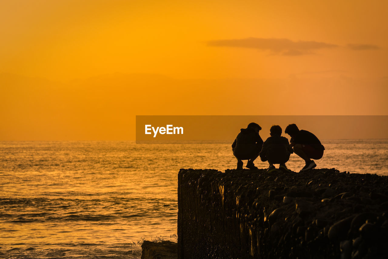 Silhouette of people crouching on rock by sea against sky during sunset