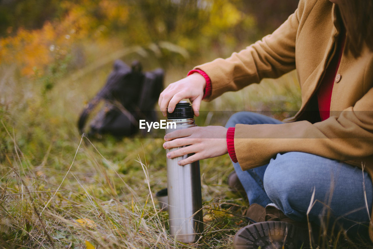 Low section of woman holding insulated drink container while sitting on field during autumn