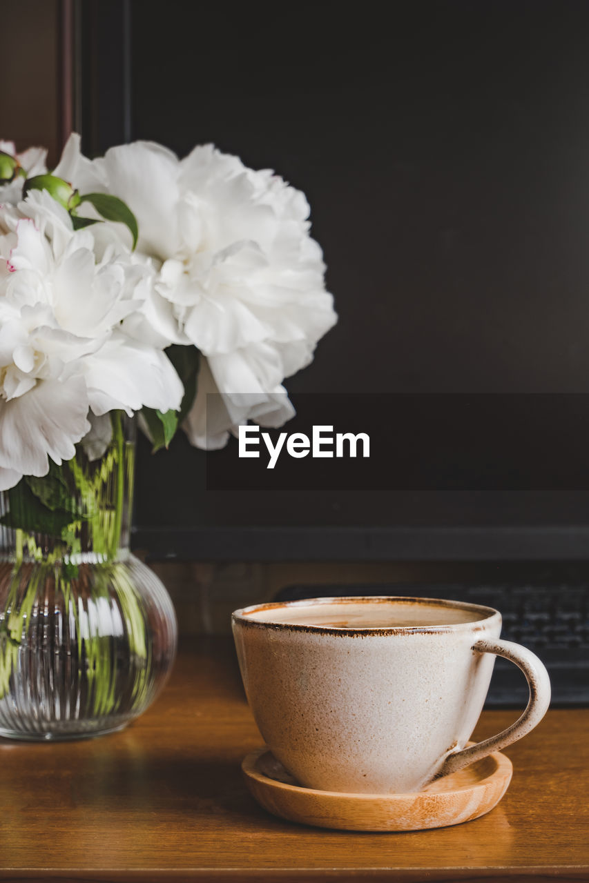 Cozy home workplace. stylish cup coffee, small glass vase with white peonies near computer monitor