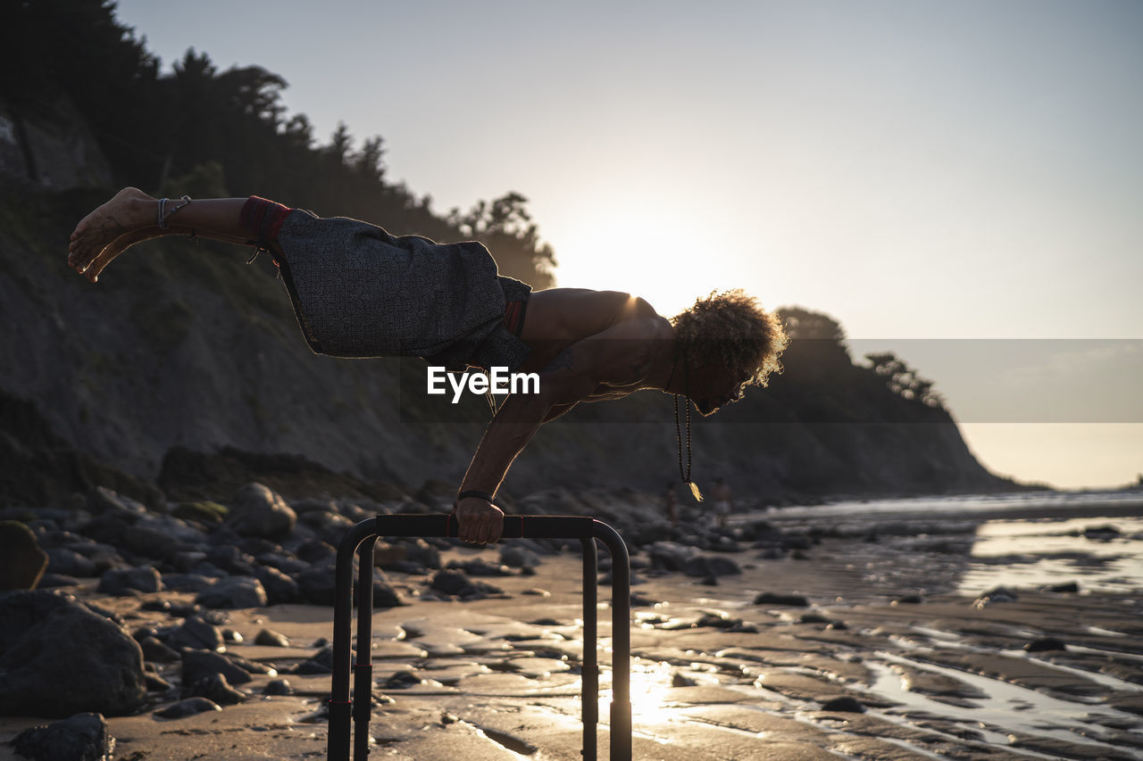 Shirtless young man exercising on parallel bars at beach against sky