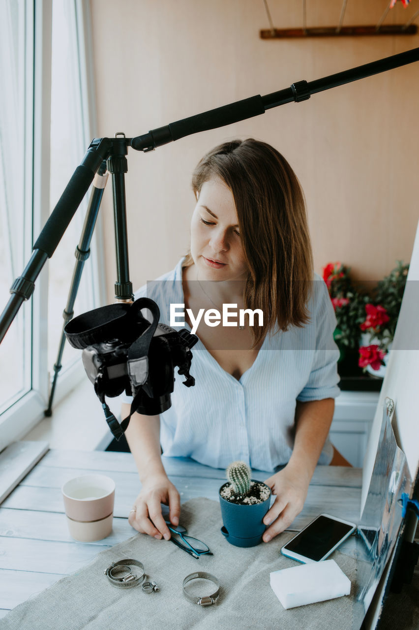 Young woman photographer in blue shirt shoots flatlay on camera tripod at home. remote working place