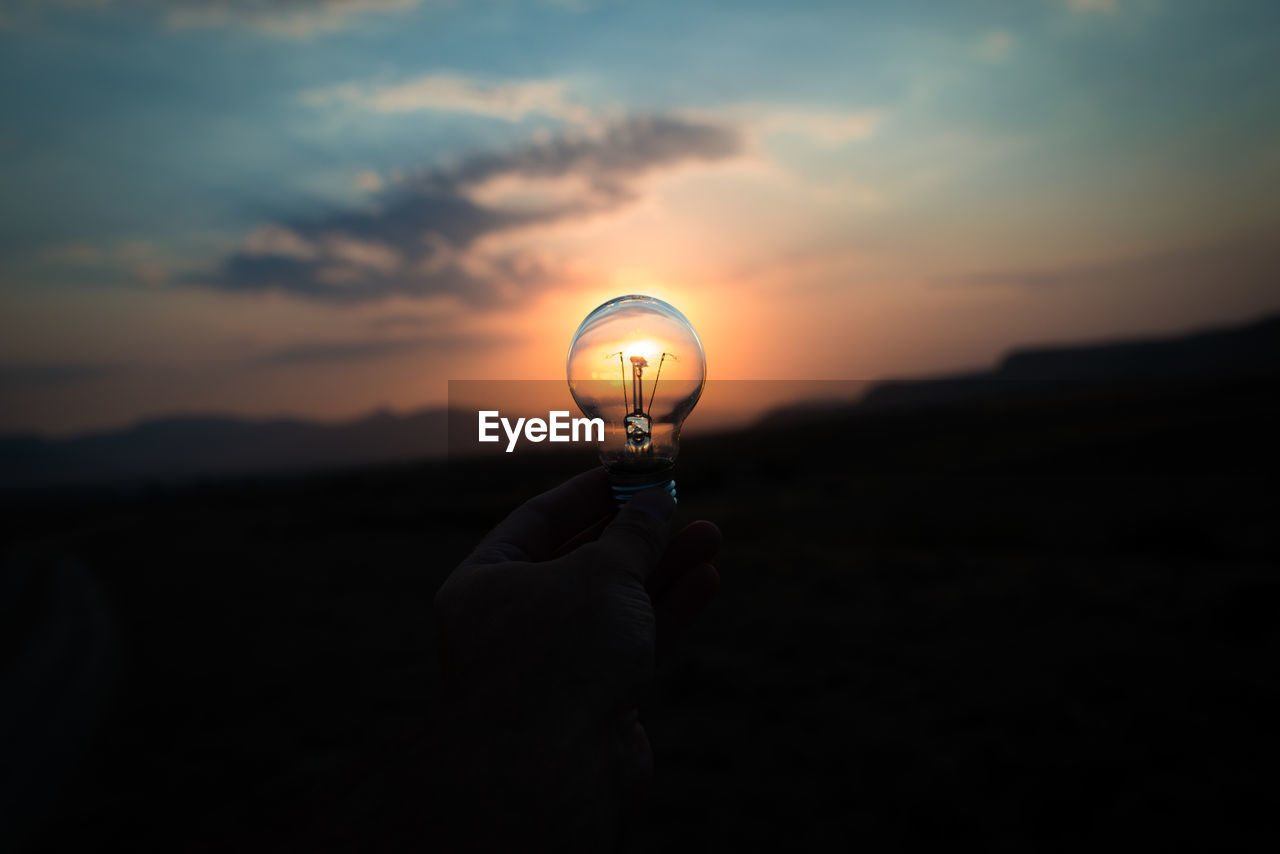 Silhouette hand holding light bulb against sky during sunset