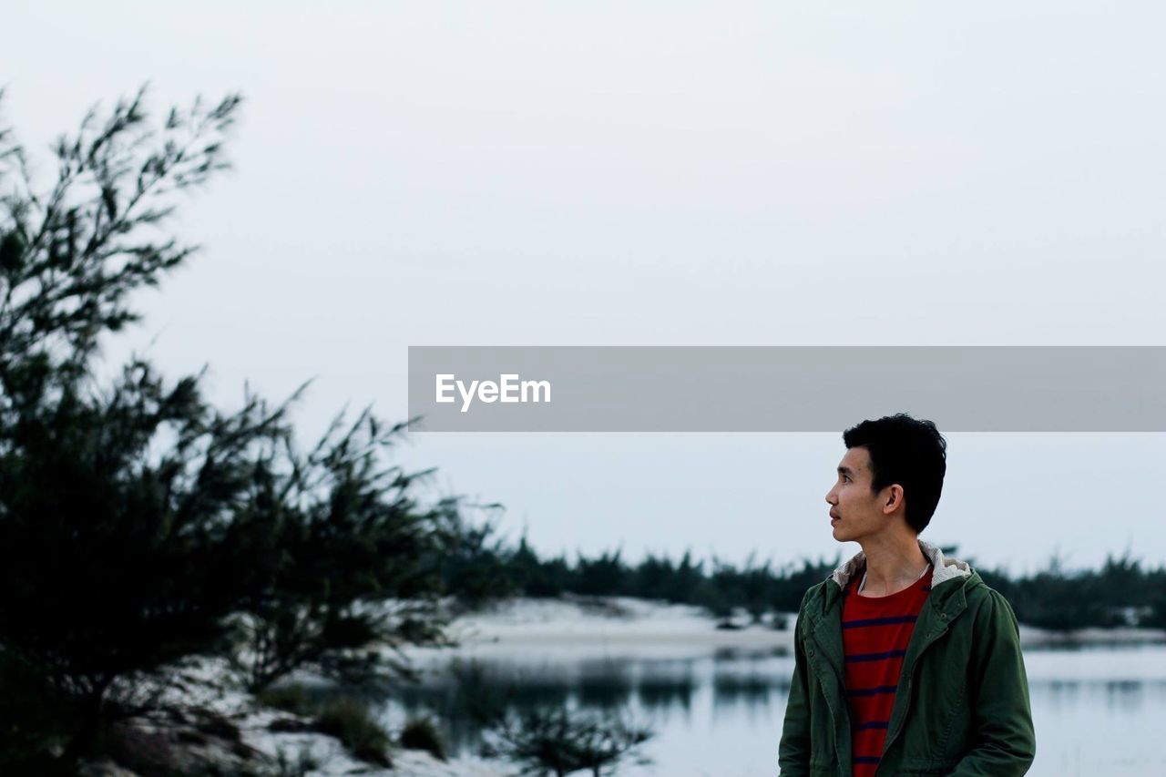 Young man looking away while standing against clear sky