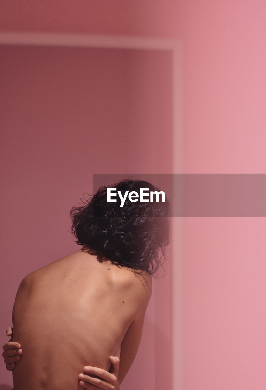 Rear view of shirtless sensuous woman standing against pink wall
