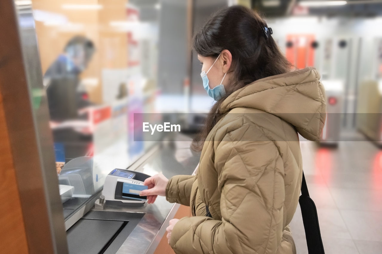 Young woman in a mask buys a subway ticket and pays with credit card contactless