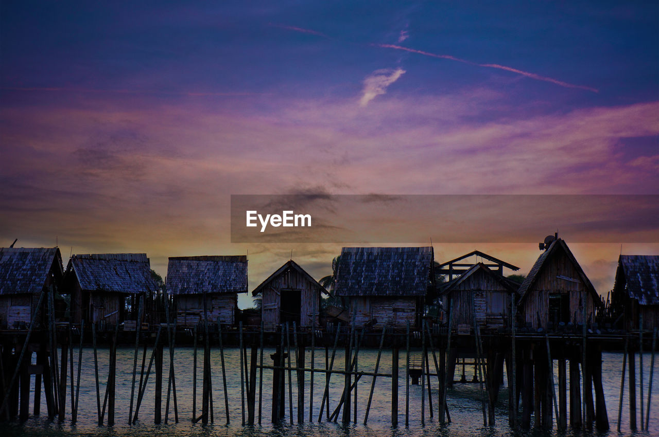 Stilt houses in sea against sky during sunset