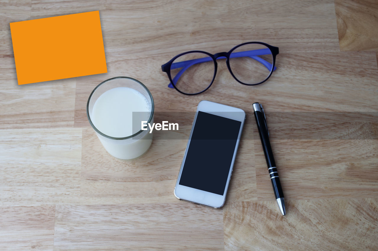 High angle view of smart phone with eyeglasses pen and milk in glass on table