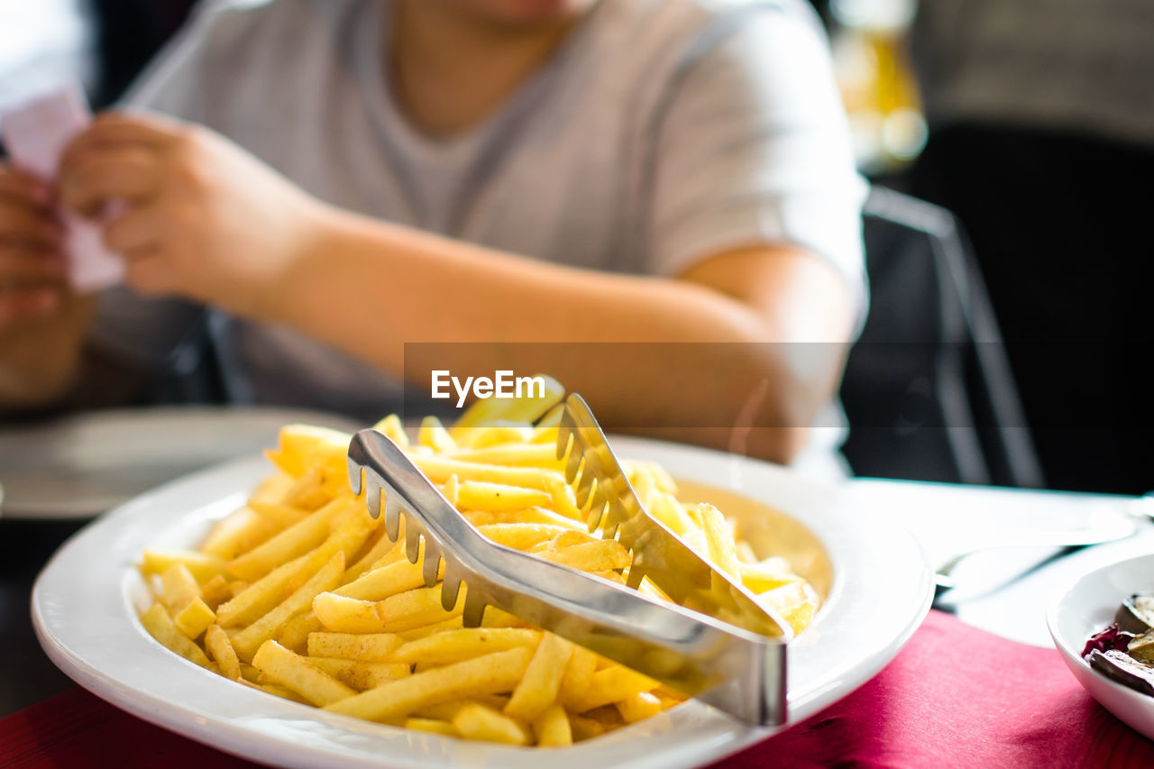 Serving tongs on french fries in plate with man sitting at table in restaurant