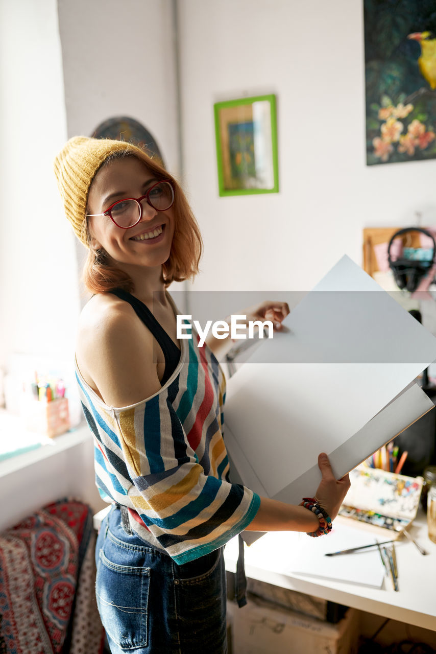 Side view of content female painter in funky outfit standing with art supplies in studio and looking at camera