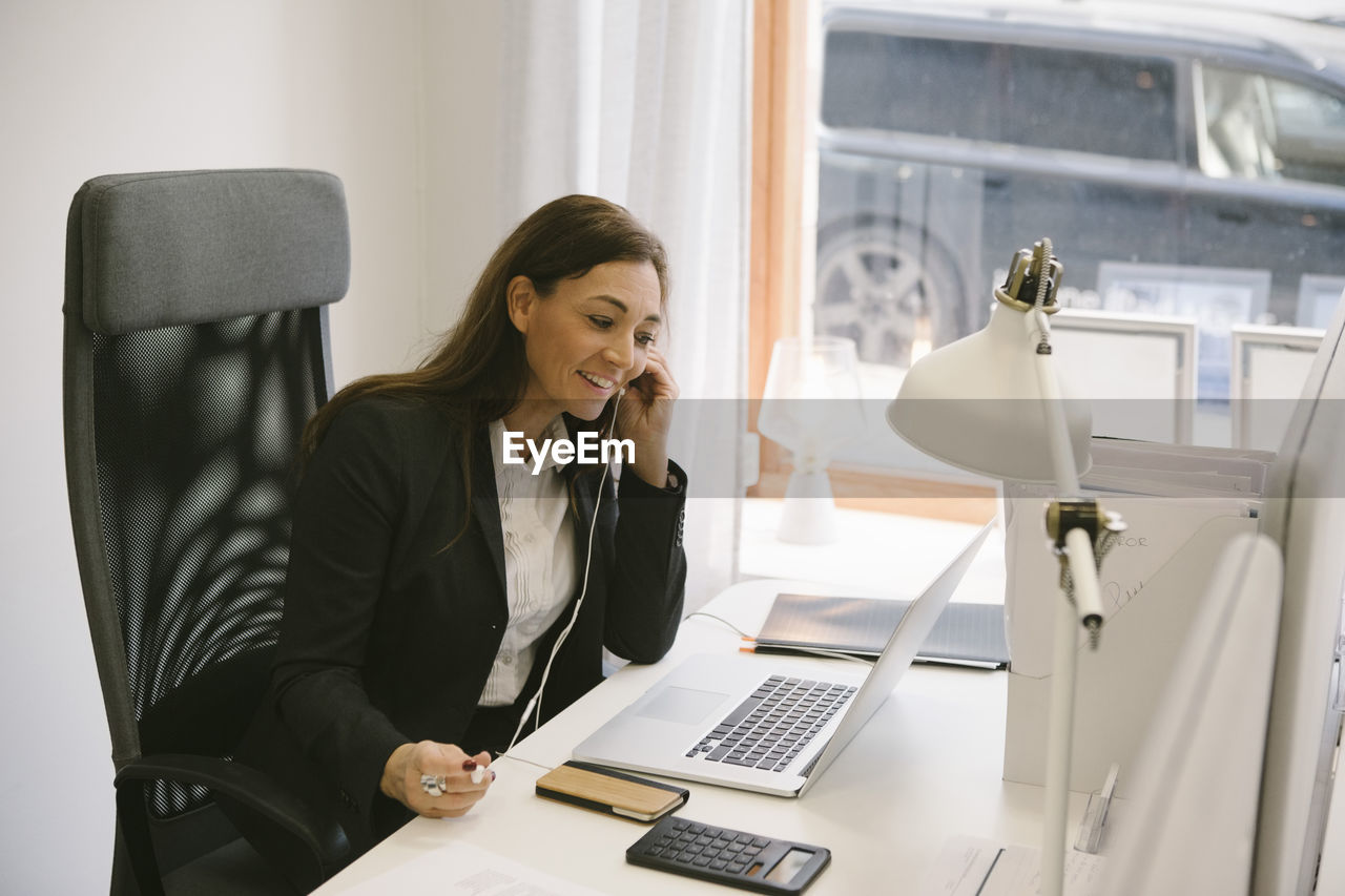 Happy realtor listening to headphones while looking at laptop in office