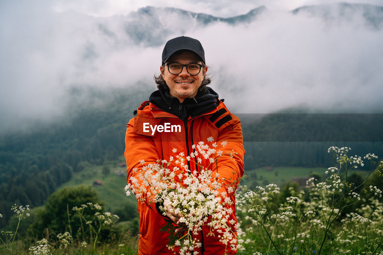 Smiling man standing on field and holding wildflowers  against misty mountain in summer