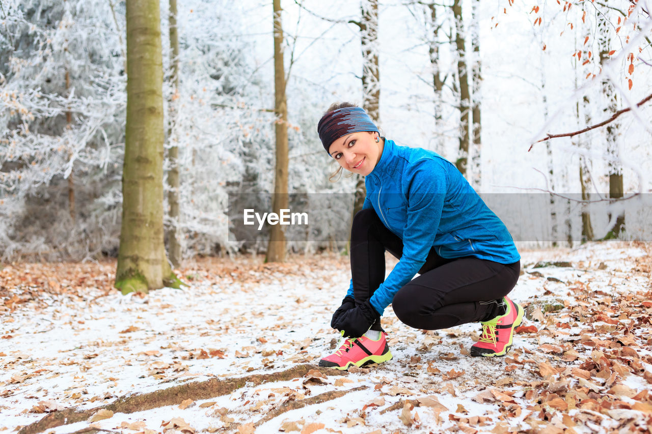 Portrait of mid adult woman tying shoelace while crouching on snow covered field in forest