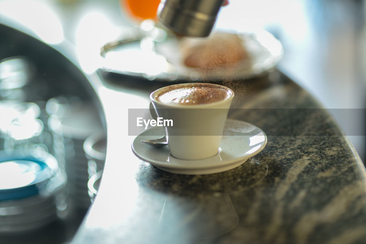 Pouring cocoa on cappuccino in a bar counter top