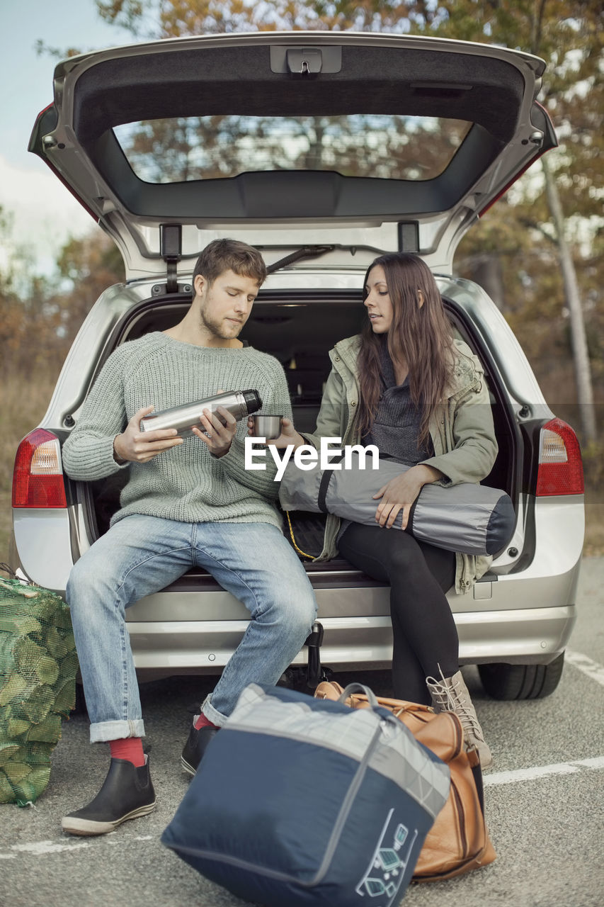 Man pouring coffee for woman while sitting at car trunk during camping