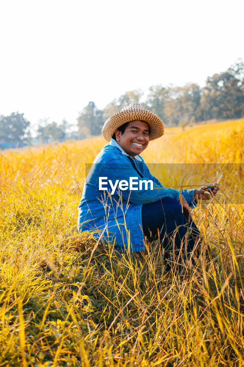 agriculture, field, one person, plant, rural scene, adult, landscape, nature, meadow, yellow, hat, land, clothing, crop, sky, smiling, rapeseed, flower, women, happiness, farm, grass, leisure activity, day, rural area, prairie, lifestyles, emotion, beauty in nature, environment, portrait, growth, copy space, tranquility, outdoors, sunlight, relaxation, autumn, senior adult, casual clothing, three quarter length, farmer, tranquil scene, grassland, person, seniors, non-urban scene, summer, clear sky, plain, flowering plant, enjoyment, looking at camera, harvesting, sunny, female, looking, sitting