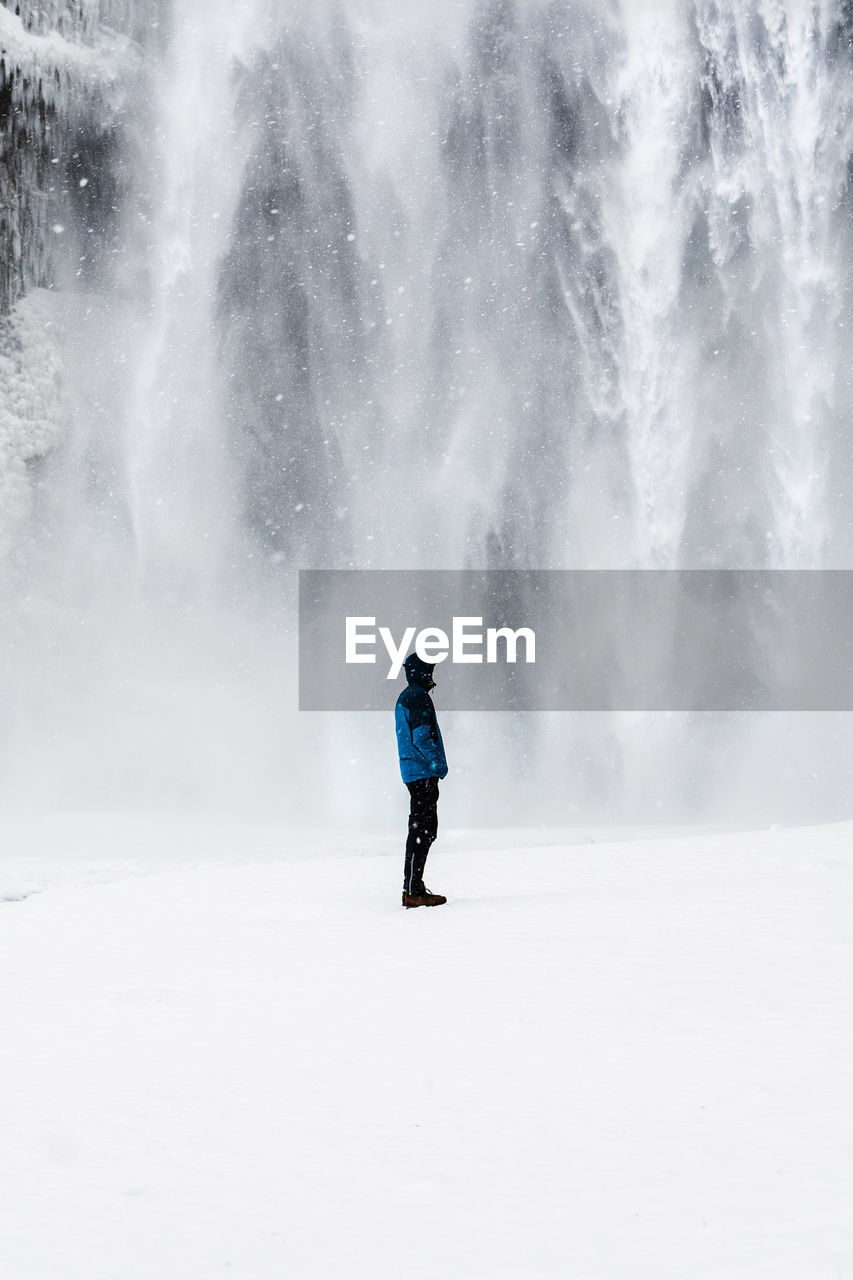 Side view of man standing on snow covered field against waterfall