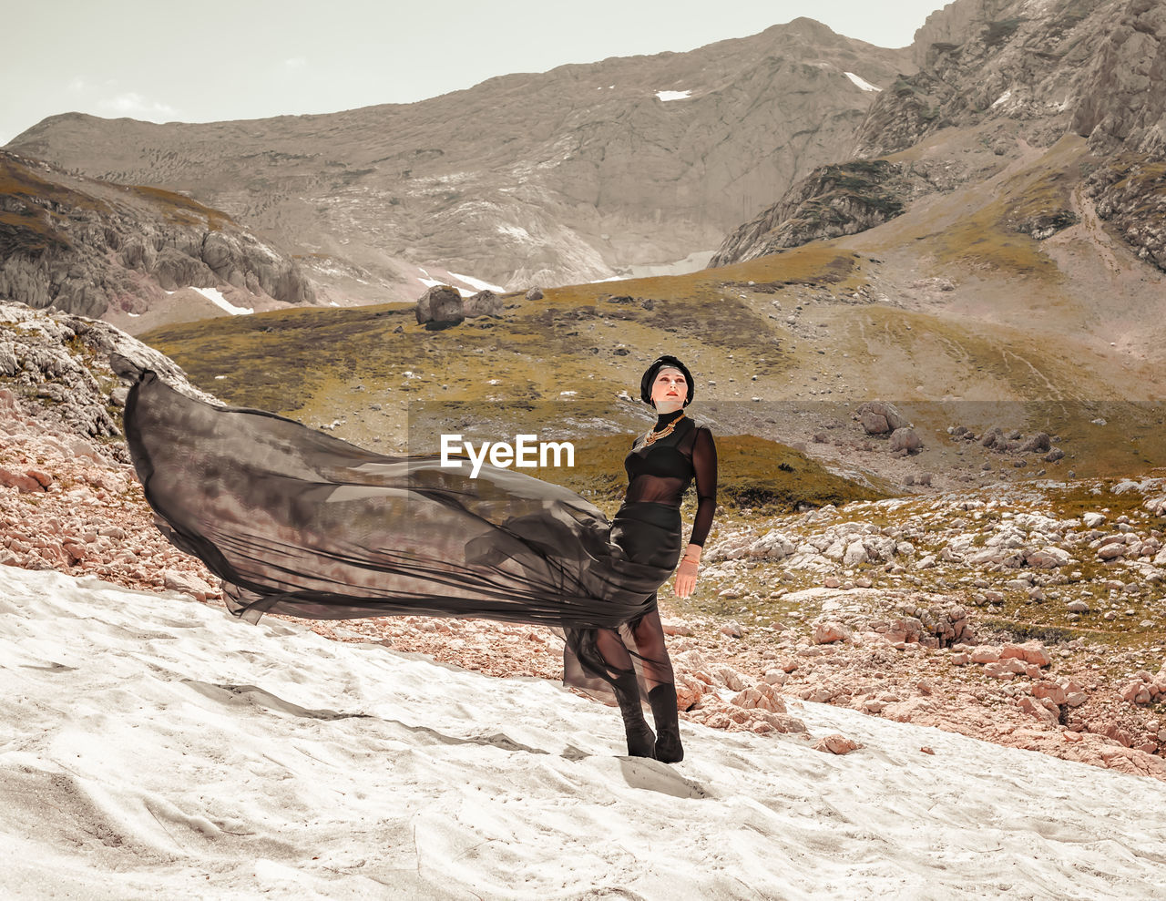A stylish determined woman in a black dress fluttering in the wind stands on the snow glacier