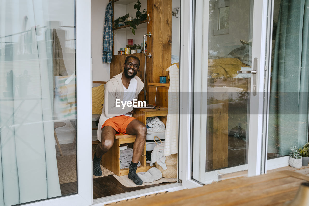 Portrait of happy man with laptop sitting on wooden seat seen through doorway