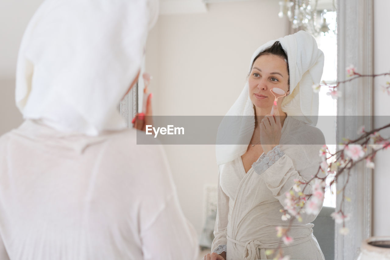 Portrait woman wrapped in white towel and in bathrobe looks at reflection