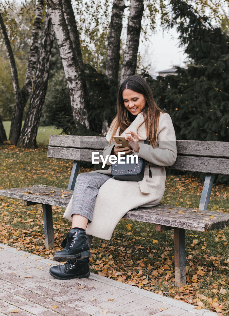 Full length of woman using phone while sitting on bench at park