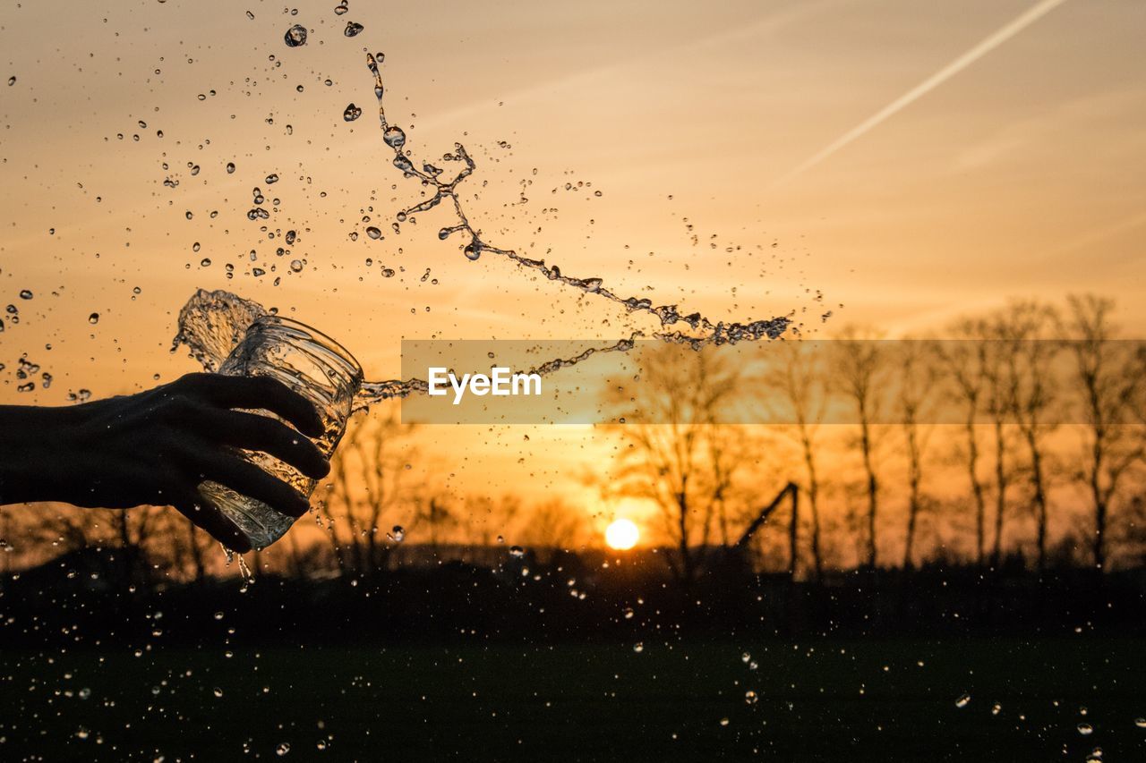 Cropped hand splashing water against sky during sunset