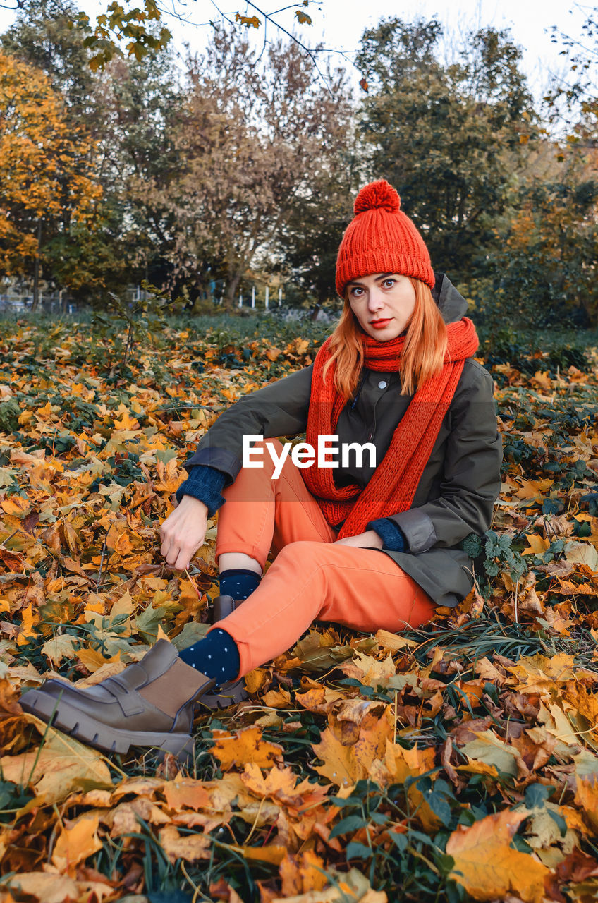 Autumn portrait of cute girl in orange warm knitted hat and orange scarf sitting in fallen leafs