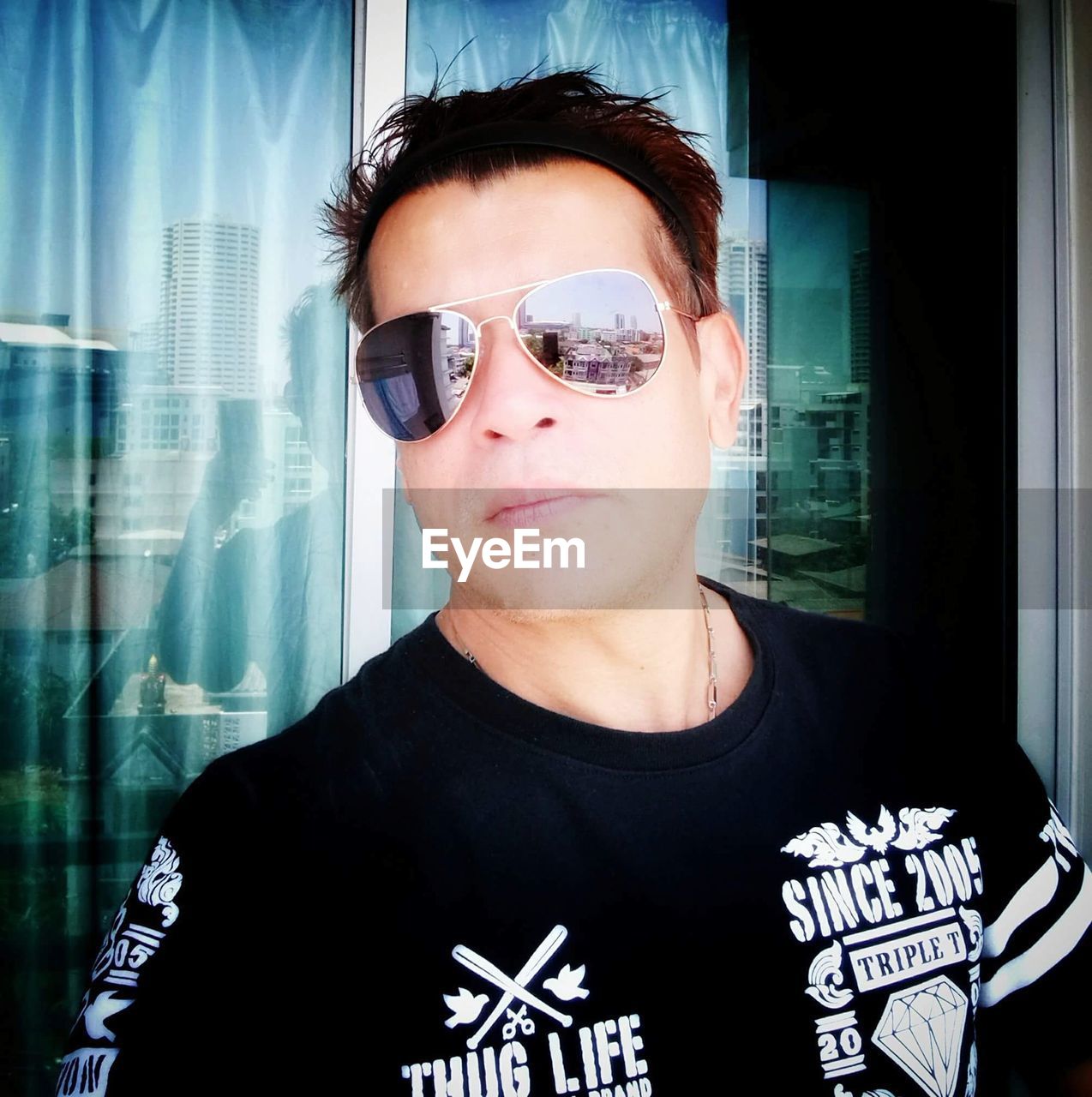 CLOSE-UP PORTRAIT OF YOUNG MAN WEARING SUNGLASSES STANDING AGAINST WINDOW