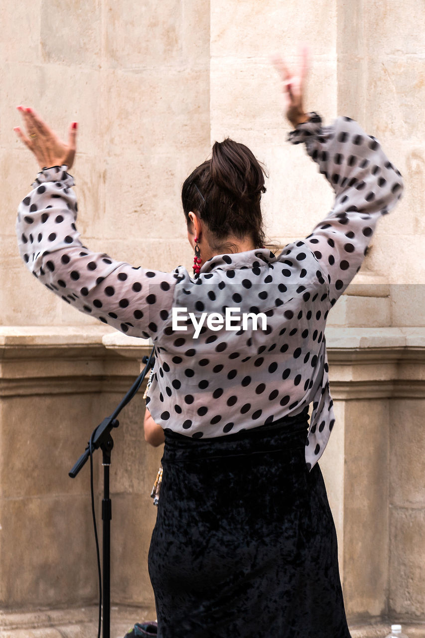 An averted woman in a dotted blouse raises her hands to sing or dance in the street
