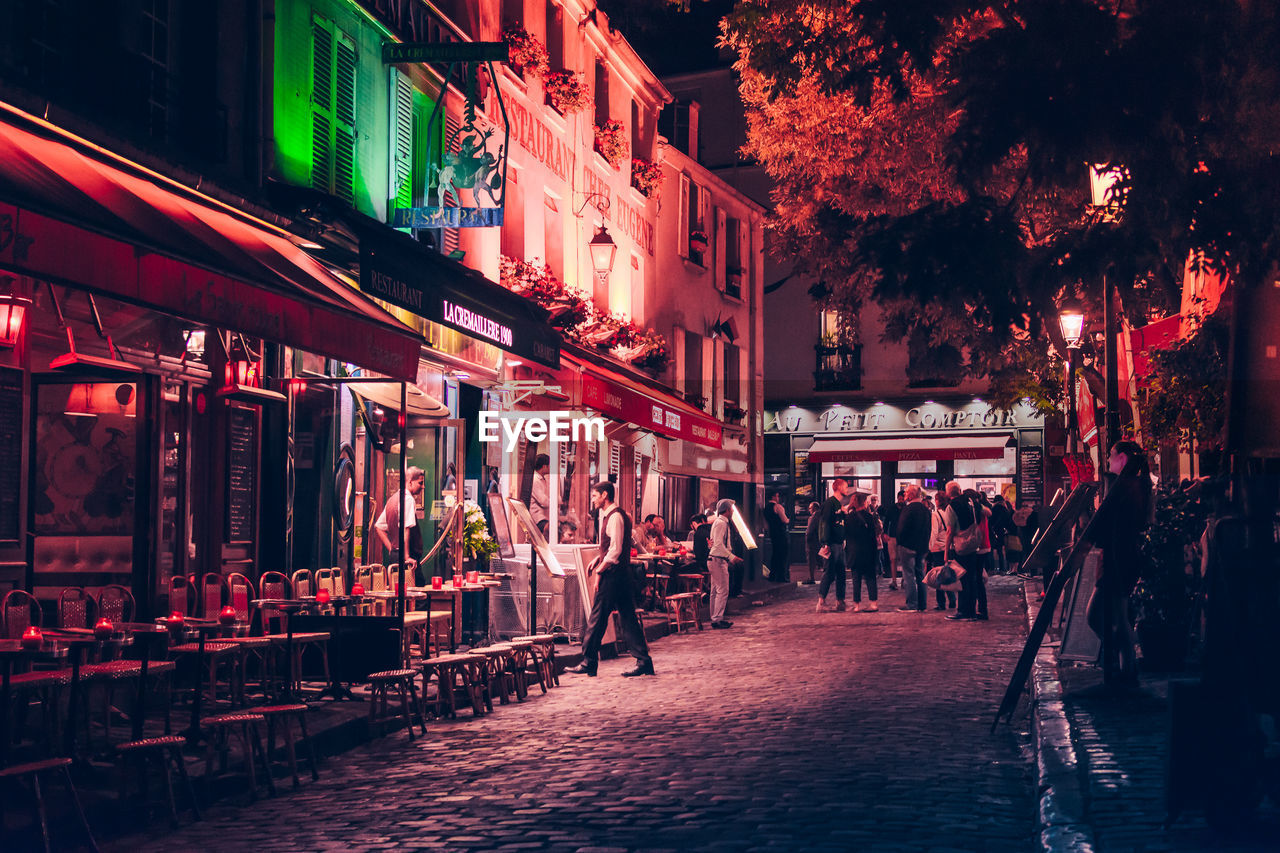 PEOPLE WALKING ON STREET AMIDST BUILDINGS AT NIGHT