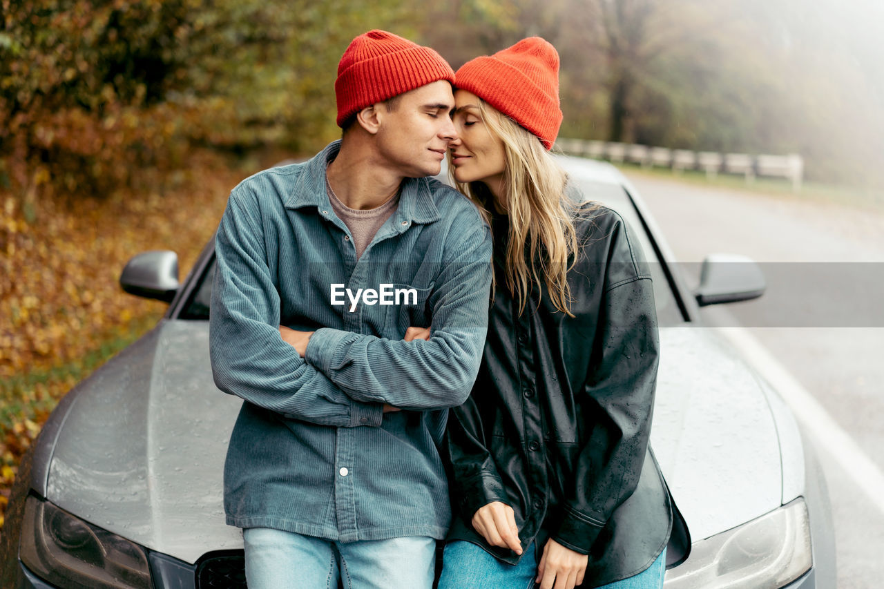 A young man and woman embrace tenderly while sitting on the bonnet of a car.