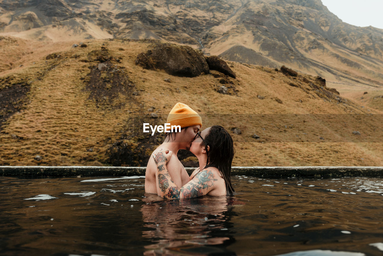 Side view of young tattooed hipsters kissing and hugging in water between mountains