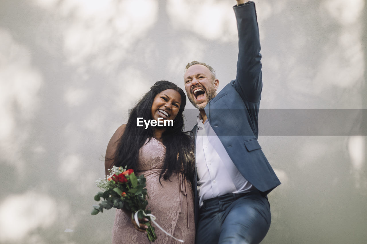 Cheerful mature groom screaming by young bride with bouquet against wall at wedding