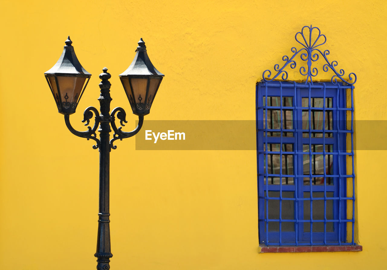 Vintage style gorgeous street lamp against yellow rough wall with a vibrant blue artistic window