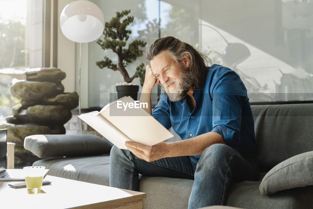 Mature man with a beard reading a book sitting on a sofa in the living room of his house. learning concept
